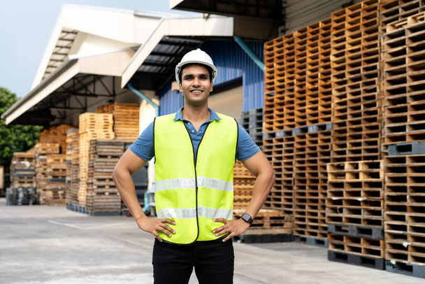 Portrait of young Indian worker working in logistic industry outdoor in front of factory warehouse. Smiling happy man with hard hat looking at camera hands on hip at depot - Foto, Imagen
