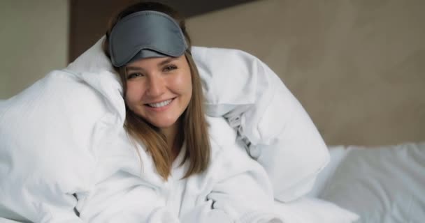 Улыбающаяся женщина в маске для сна лежит под одеялом на кровати крупным планом - Кадры, видео