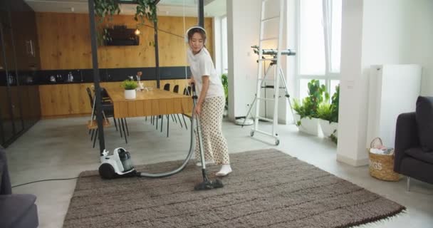 Asian woman listening music in white headphones pretending playing guitar while vacuum cleaning carpet floor in living room at home. Concept of housekeeping, family cleaning house, lifestyle. - Πλάνα, βίντεο