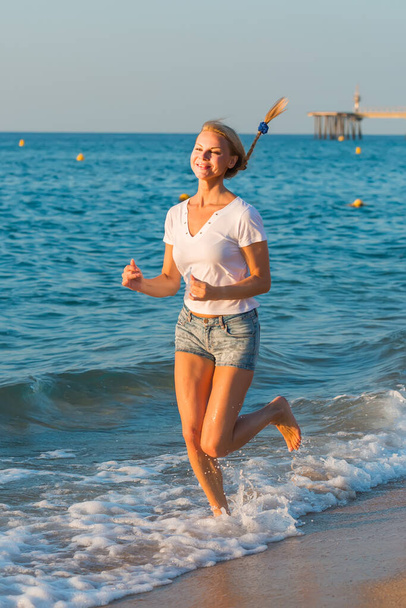 Smiling adult woman in white T-shirt is jogging on the beach - Foto, immagini