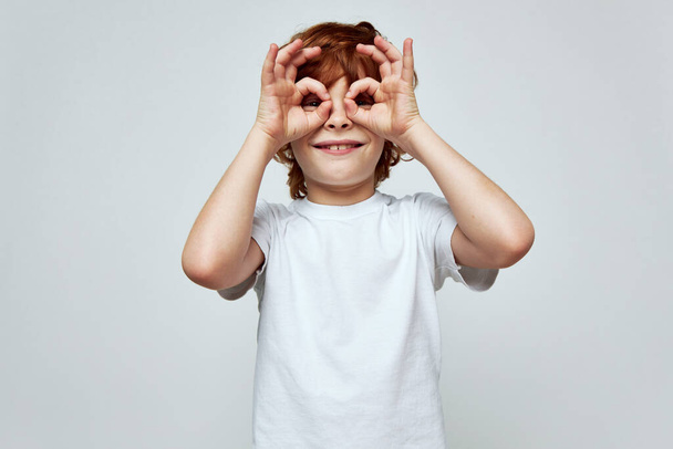alegre sorridente ruiva menino segurando as mãos perto de seus olhos na forma de uma máscara branca t-shirt  - Foto, Imagem