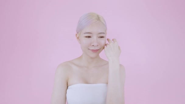 Азійка, яка м'яко торкається шкіри обличчя, закохується в мить доброго здоров'я шкіри після прибирання. Сучасна блондинка і рожеве волосся красива жіноча модель на ізольованому рожевому фоні вдягають сексуальний білий бюстгальтер - Кадри, відео