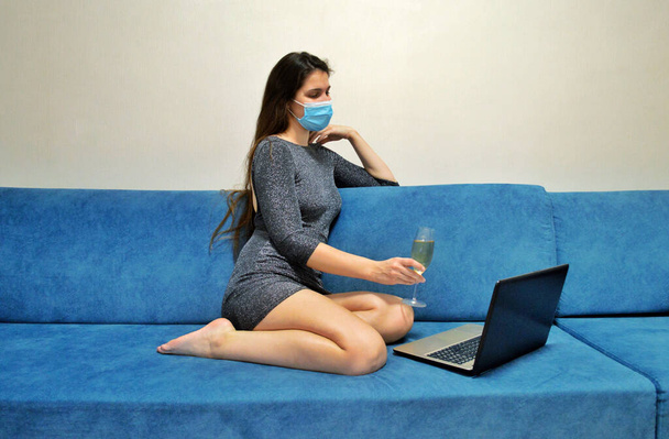 κορίτσι κάθεται σε μάσκα στον καναπέ με σαμπάνια στα χέρια της μπροστά από ένα φορητό υπολογιστή - Φωτογραφία, εικόνα