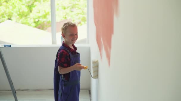 Хлопець європейського походження малює стіни будинку роликом. вдягнені в комбінезон - Кадри, відео