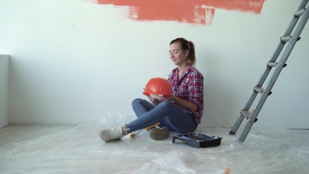 Μια γυναίκα κάθεται στο πάτωμα ενός σπιτιού, βάζει ένα πορτοκαλί προστατευτικό κράνος στο κεφάλι της. Σχέδιο ανακαίνισης σπιτιού - Πλάνα, βίντεο