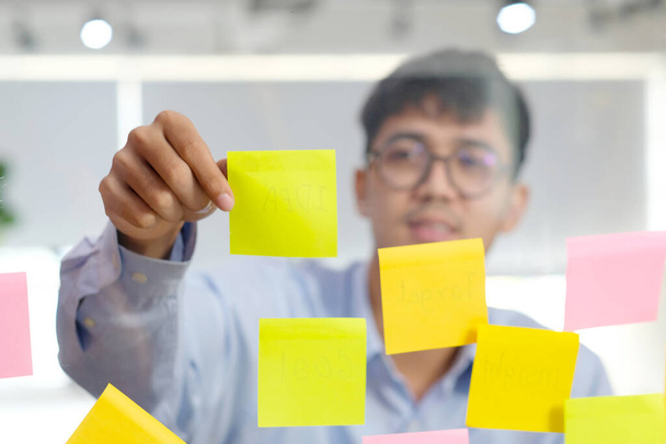 Jovem asiático homem ler nota pegajosa na parede de vidro no escritório, negócios brainstorming ideias criativas, estilo de vida de escritório, sucesso no conceito de negócios - Foto, Imagem