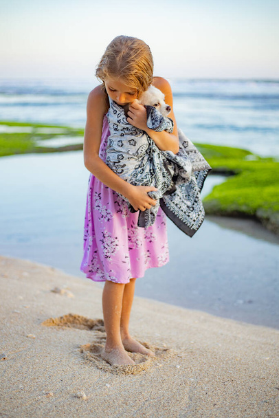 Jolie petite fille tenant petit chien chihuahua blanc avec queue rose sur la plage. Prends soin de l'animal. Chien couvert d'écharpe. Passez du temps dehors. Concept d'enfance. Plage de Melasti, Bali, Indonésie - Photo, image