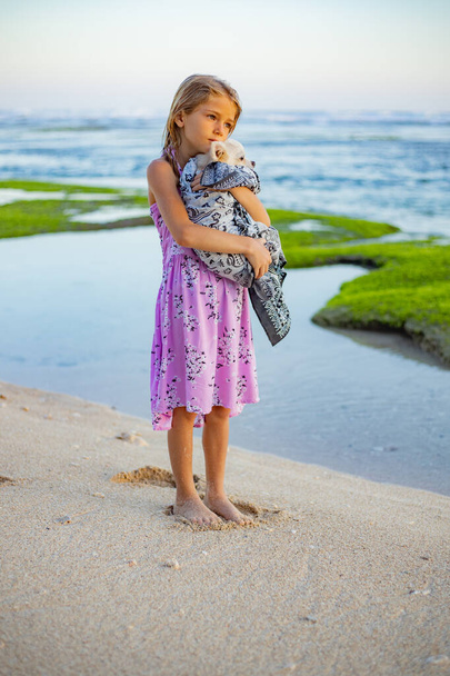 Χαριτωμένο κοριτσάκι κρατώντας μικρό λευκό chihuahua σκυλί με ροζ ουρά στην παραλία. Φρόντισε το κατοικίδιο. Σκύλος καλυμμένος με μαντήλι. Πέρνα λίγο χρόνο έξω. Παιδική έννοια. Παραλία Μελαστή, Μπαλί, Ινδονησία - Φωτογραφία, εικόνα