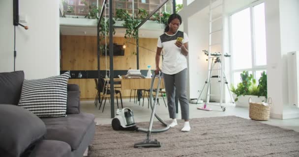 Joyful african american woman hoovering floor in modern house with vacuum cleaner and texting chatting with friends on social media using smartphone. Concept housekeeping, cleaning house, lifestyle. - Πλάνα, βίντεο