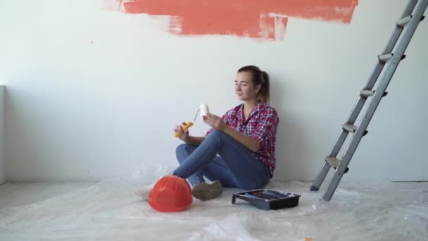 Eine Frau sitzt auf dem Fußboden des Hauses und hält eine Walze in der Hand, um die Wände zu streichen. Sanierungskonzept für Eigenheime - Filmmaterial, Video