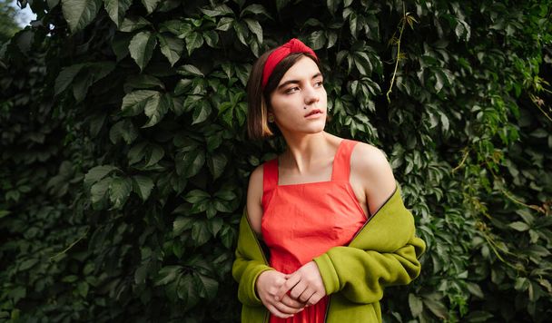 Portrait of beautiful young woman staying in green park garden trees  natural background. Red dress and accessories green stylish jacket. Fashion style. Romantic mood photo. pensively looks aside.  - Zdjęcie, obraz