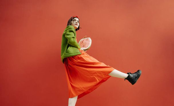 Watermelon cat walk conceptual fashion style. girl holding half of fruit.  Red dress and background and green jacket. leg wit black boot raised. Looking curious to the side. Beautiful teenager girl  - Фото, изображение