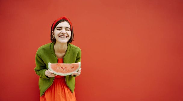 Adorable laughing brunet teen girl in green jacket and red dress. Hair accessories. Holding slice of watermelon. Funny joke. sense of humor concept. Looking to the camera teeth smile. Street style  - 写真・画像