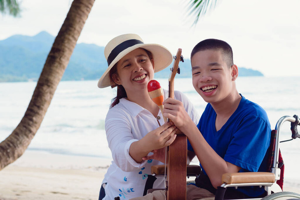 Asian special child on wheelchair is singing, playing ukulele happily on the beach with parent,Natural sea beach background,Life in the education age of disabled children,Happy disability kid concept. - Photo, Image