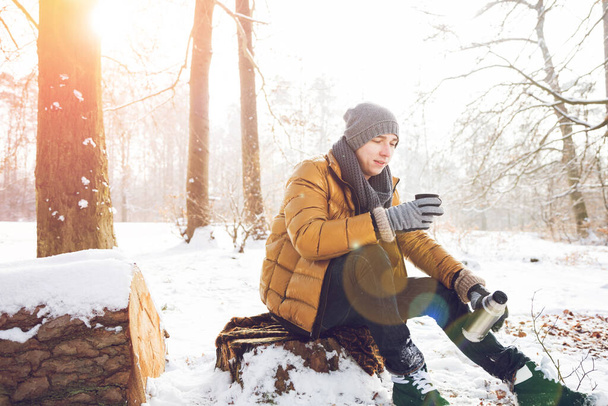 young man pouring hot beverage from thermos and sitting in winter forest - Foto, Bild