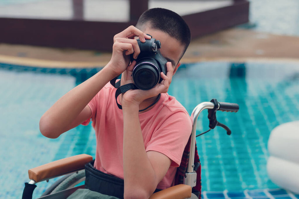 Camera in hands of Asian special child on wheelchair take photo at swimming pool in front of sea beach,Boy happiness in holidays with family time on the travel,Lifestyle of happy disabled kid concept. - Photo, Image