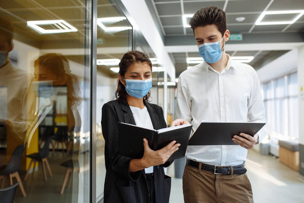 Az üzletemberek munkamegbeszéléseket folytatnak a koronavírus világjárvány idején. Férfi és nő védő orvosi maszkot visel, üzletről beszélgetnek az iroda folyosóján. Munkahelyi egészségvédelem - Fotó, kép