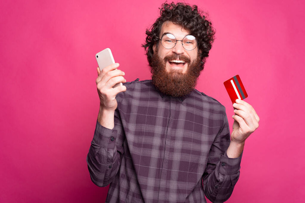 A bearded excited man is holding a credit card and his phone near a pink wall - Foto, afbeelding