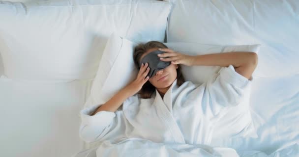 Lächelnde Dame mit Maske erwacht auf großem bequemen Bett liegend - Filmmaterial, Video