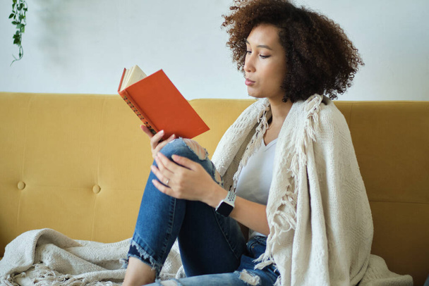 Прекрасна молода афроамериканська жінка, яка читає книжку, лежачи зручно на спині на дивані у вітальні в джинсах. - Фото, зображення