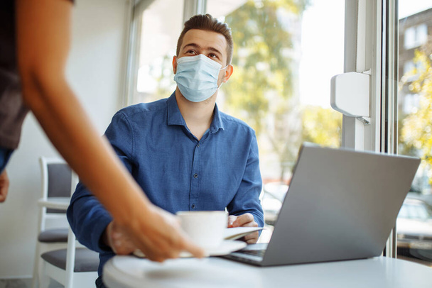 Waiter brings a cup of coffee to a young businessman wearing a sterile medical mask working from a cafe remotely. Distant business concept during coronavirus pandemic - Photo, Image