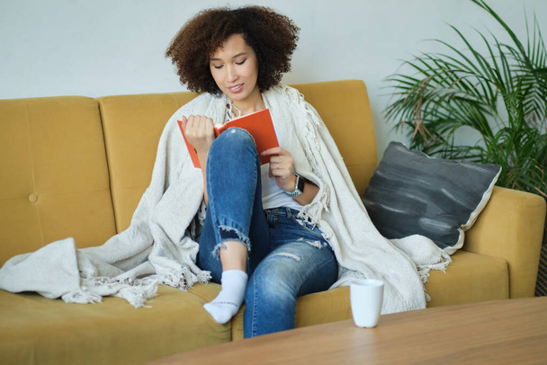 Hermosa joven afroamericana leyendo un libro acostada cómodamente sobre su espalda en un sofá en la sala de estar en sus pantalones vaqueros - Foto, imagen