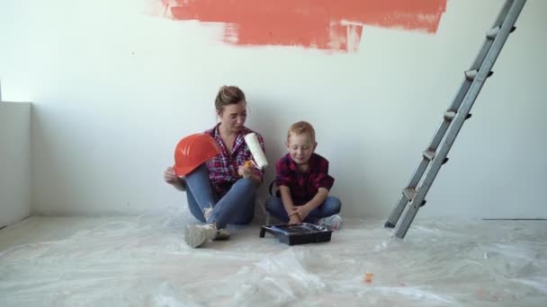 Η μαμά και ο γιος κάθονται στο πάτωμα και αναπαύονται αφού βάψουν τους τοίχους. Σχέδιο ανακαίνισης σπιτιού - Πλάνα, βίντεο