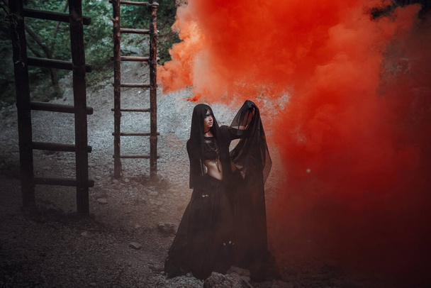 Φανταστική μάγισσα τρόμου με μαύρο φόρεμα. Γοτθικό μαγικό πορτρέτο. Κακό πρόσωπο μακιγιάζ Τέχνη τρομακτικό, Μάγος βαριά ξόρκι κόκκινο καπνό. - Φωτογραφία, εικόνα