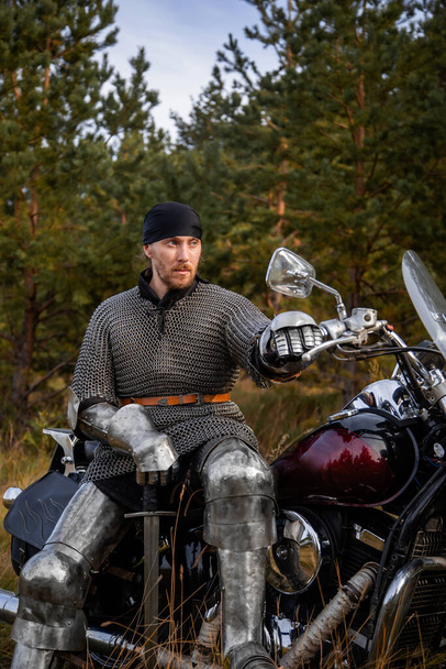 A medieval warrior in armor sits on a motorcycle with a sword in his hands on the background of the forest. Knight biker concept. - Fotó, kép