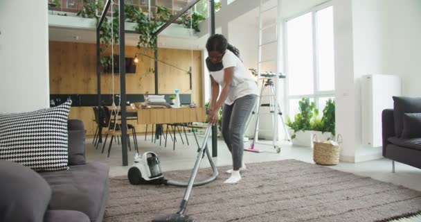 Joyful african american woman using modern vacuum cleaning carpet floor in a bright cozy living room at home while speaking on phone. Concept of housekeeping, cleaning house, lifestyle. - Filmagem, Vídeo