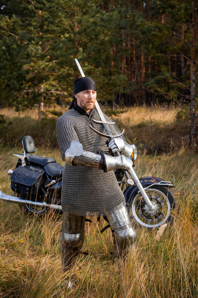 A medieval warrior in armor with a two-handed sword in his hands stands next to a motorcycle against the backdrop of a forest. Knight biker concept. - Photo, image
