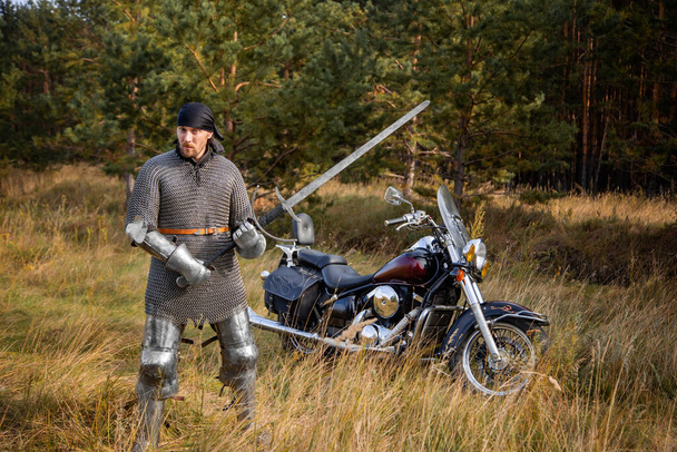 A medieval warrior in armor with a two-handed sword in his hands stands next to a motorcycle against the backdrop of a forest. Knight biker concept. - Photo, Image