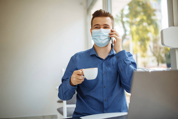 医療用の滅菌マスクを着用した若いビジネスマンがカフェから携帯電話の主要なビジネスについて話し合い、コロナウイルスパンデミックの検疫中にリモートで働いています。健康安全コンセプト - 写真・画像