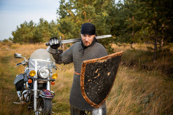 A medieval knight in chainmail with a shield and a sword in his hands stands against the backdrop of a motorcycle and a forest. - Fotoğraf, Görsel