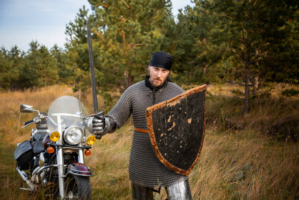A medieval knight in chainmail with a shield and a sword in his hands stands against the backdrop of a motorcycle and a forest. - Foto, immagini