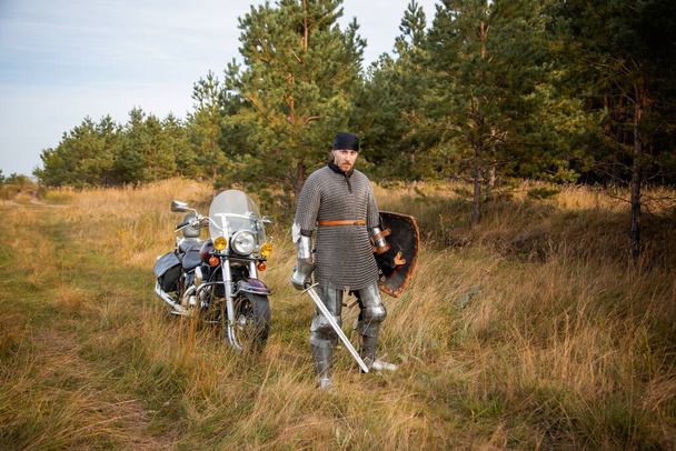 A medieval knight in chainmail with a shield and a sword in his hands stands against the backdrop of a motorcycle and a forest. - Photo, image