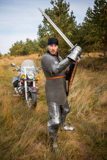 A medieval knight in chainmail with a shield and a sword in his hands stands against the backdrop of a motorcycle and a forest. - Foto, afbeelding
