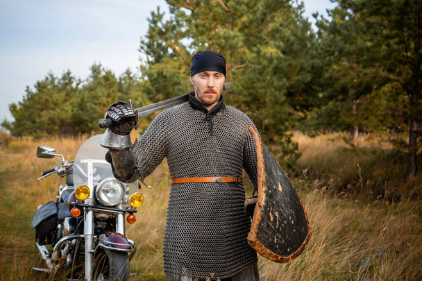 A medieval knight in chainmail with a shield and a sword in his hands stands against the backdrop of a motorcycle and a forest. - Φωτογραφία, εικόνα
