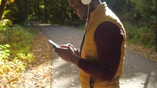 Concepto de escuchar música y podcasts. Sonriendo joven afroamericano hombre escucha música en un parque de otoño - Imágenes, Vídeo