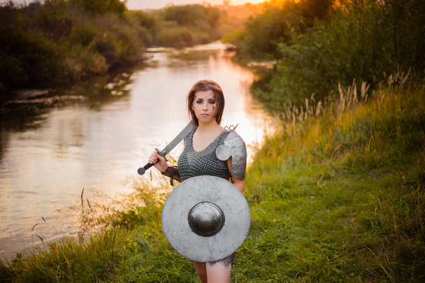 Medieval fantasy female warrior with war paint on her face, in chain mail armor, with a sword and shield in her hands, poses standing against the backdrop of a sunset, river and forest. - Fotografie, Obrázek