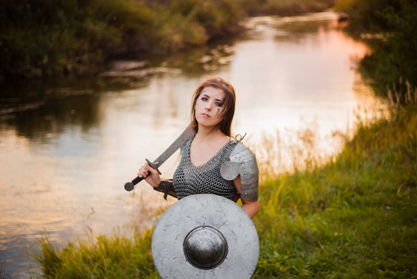 Medieval fantasy female warrior with war paint on her face, in chain mail armor, with a sword and shield in her hands, poses standing against the backdrop of a sunset, river and forest. - Valokuva, kuva