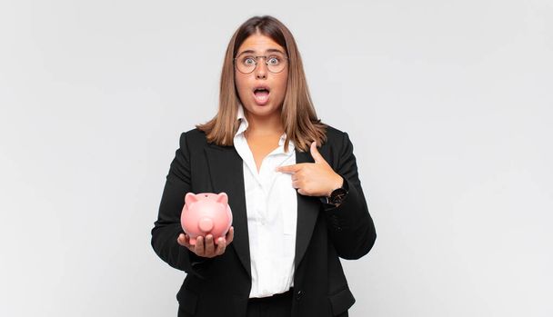 young woman with a piggy bank looking shocked and surprised with mouth wide open, pointing to self - Photo, Image