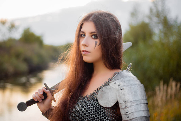 Portrait of a medieval woman warrior in chain mail with a sword and shield in her hands against the backdrop of the river and sunset. Combat makeup, clear skin, retouching. - Foto, Bild