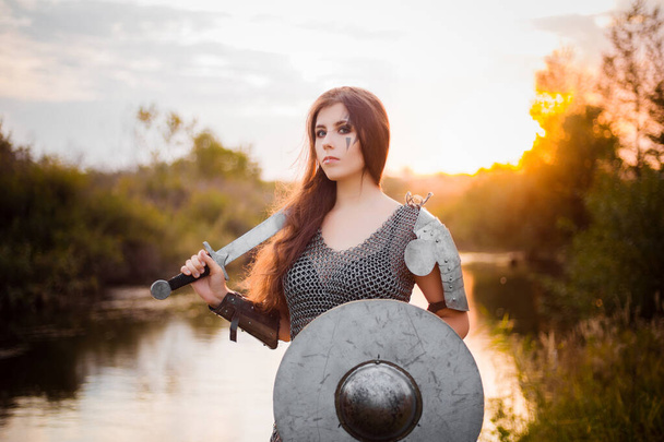Portrait of a medieval woman warrior in chain mail with a sword and shield in her hands against the backdrop of the river and sunset. Combat makeup, clear skin, retouching. - Foto, imagen