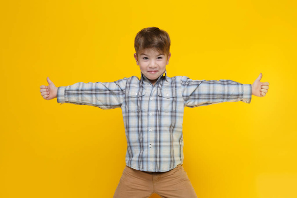 Cute little boy in a plaid shirt shows a thumb raised to the top on both arms outstretched to the sides isolated on a yellow background. - Photo, image