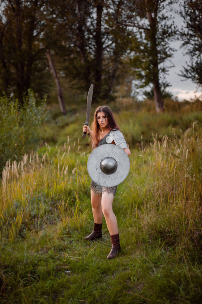 A medieval female warrior dressed in chain mail with a sword and shield in her hands poses against the background of a forest. Fantasy costume, combat makeup. - Valokuva, kuva