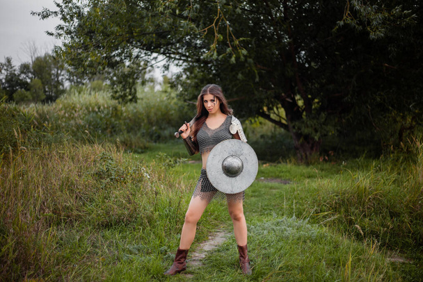 A medieval female warrior dressed in chain mail with a sword and shield in her hands poses against the background of a forest. Fantasy costume, combat makeup. - Foto, Imagen
