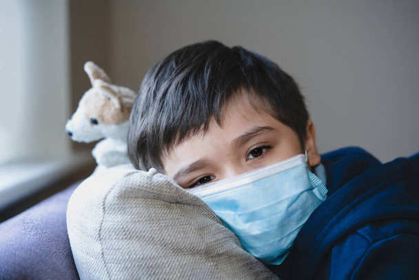 Sick child wearing a protective mask, ill kid in a medical face mask lying head on sofa with sad face,Upset boy sitting on couch looking deep in thought, Patient isolated in house to prevent infection - Fotó, kép