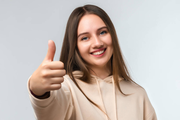 Счастливая молодая женщина с длинными каштановыми волосами, улыбающаяся и показывающая большой палец вверх, одетая в повседневную одежду. Мне это нравится. Понятие человеческих эмоций - Фото, изображение