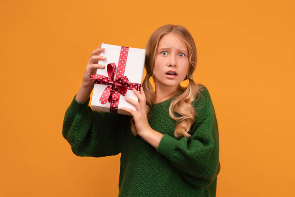 Изображение очаровательной блондинки 12-14 лет в тёплом зелёном свитере, держащей настоящую коробку с красным бантом. Студийный снимок, жёлтый фон, изолирован. Концепция новогодних праздников - Фото, изображение